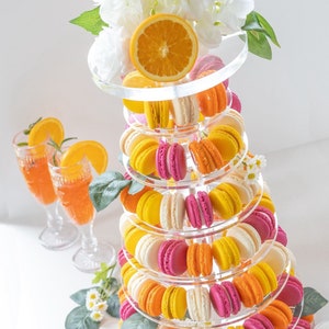 May include: A tiered acrylic stand with a white floral topper, holding a variety of colourful macarons. The macarons are arranged in a circular pattern on each tier, with alternating colours of pink, yellow, and orange. The stand is perfect for displaying desserts at a party or special event.