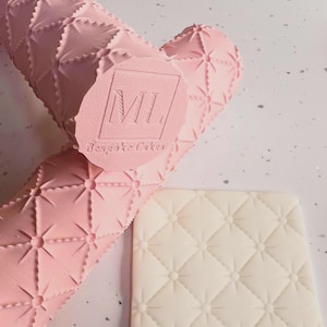 May include: A pink rolling pin and a square fondant stamp with a quilted pattern. The rolling pin has a repeating diamond pattern with starburst accents. The stamp is off-white and features a similar quilted design. The rolling pin has the text "ML Bespoke Cakes".