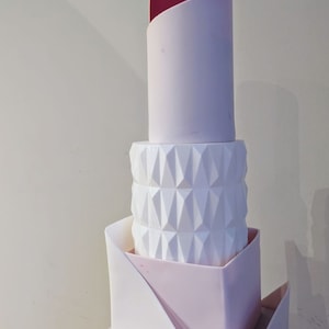 Könnte beinhalten: Eine mehrstöckige Hochzeitstorte mit geometrischem Design. Die oberste Etage ist eine weiße Torte mit einem rosa Fondant-Oberteil und einem roten Fondant-Akzent. Die zweite Etage ist eine weiße Torte mit einem geometrischen Muster. Die dritte Etage ist eine weiße Torte mit einem rosa Fondant-Oberteil. Die Torte ist mit weißem Fondant verziert.