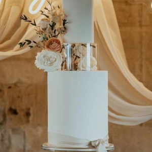 May include: A three-tiered white wedding cake with a clear glass tier in the middle. The cake is decorated with a white ribbon and a bouquet of white and peach flowers.