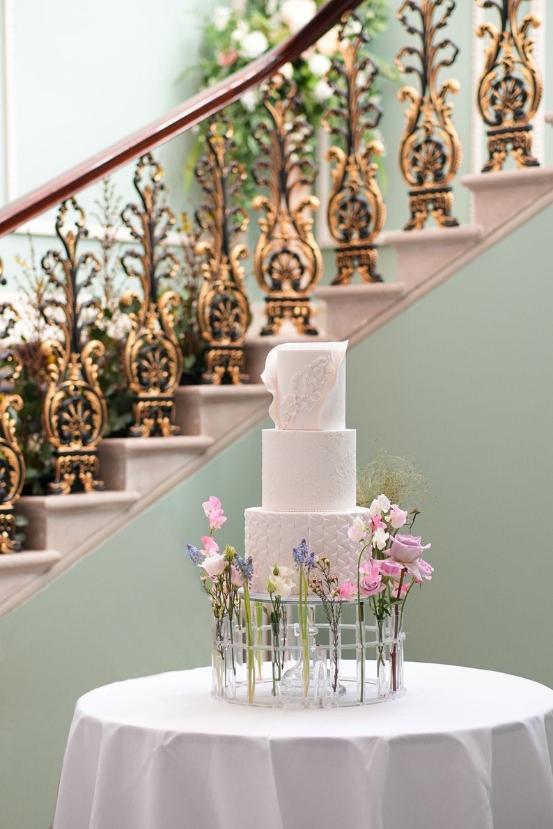 May include: A three-tiered white wedding cake with intricate designs and a delicate floral topper. The cake is displayed on a clear stand with a circular base and multiple glass tubes holding pink, purple, and blue flowers.