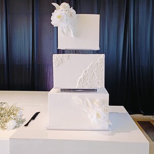 Może przedstawiać: Trzypiętrowy biały tort weselny z dekoracjami kwiatowymi. Ciasto ma kwadratowe warstwy z teksturowanymi detalami i jest ozdobione białymi kwiatami. Czarny nóż spoczywa na stole obok ciasta.