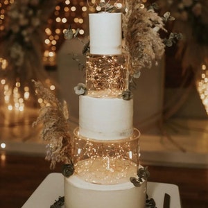 May include: A white wedding cake with four tiers, decorated with greenery and fairy lights. The top tier has a topper that says "Together".