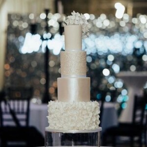 May include: A four-tier wedding cake with white frosting and a sparkling silver tier. The top tier is decorated with white sugar flowers. The cake is on a clear acrylic cake stand. Rosewood Cakes.
