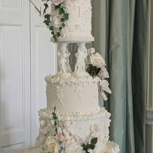 Puede incluir: Un pastel de bodas blanco de cuatro pisos decorado con flores de azúcar, querubines y detalles de encaje. El piso superior tiene las iniciales "T & B" en glaseado blanco.