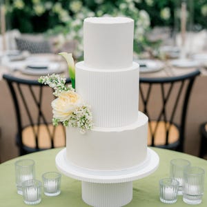 Könnte beinhalten: Eine dreistöckige weiße Hochzeitstorte mit einem schlichten, modernen Design. Die Torte ist mit weißem Frosting und einer einzelnen weißen Rose mit grünen Blättern verziert. Die Torte steht auf einem weißen Tortenständer mit geriffeltem Design. Die Torte befindet sich auf einer grünen Tischdecke mit drei klaren Glaskerzenhaltern.