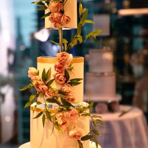 Op de afbeelding: Een vierlagen bruidstaart versierd met delicate roze rozen en groen. De taart is wit met gouden accenten en wordt gepresenteerd op een transparante standaard.