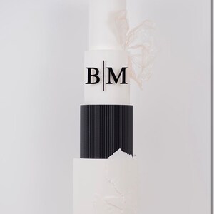 Könnte beinhalten: Eine dreistöckige Hochzeitstorte mit weißem Fondant und schwarzen Akzenten. Die oberste Etage hat eine weiße Fondantbasis mit den Initialen "B|M" in Schwarz. Die mittlere Etage ist schwarz mit vertikalen Linien. Die unterste Etage ist weiß mit einem strukturierten Design.