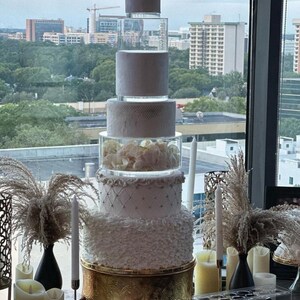 May include: A white wedding cake with multiple tiers, decorated with white frosting, lace, and flowers. The cake is on a gold stand and is in front of a window with a city view. The cake is surrounded by candles and decorative grasses.