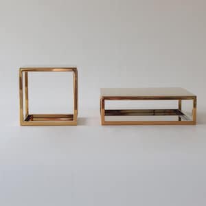 May include: Two gold-coloured metal tables. One is a cube-shaped side table, and the other is a rectangular coffee table. Both tables have a mirrored surface and a minimalist design. The tables are suitable for a living room or other home decor.