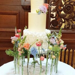 May include: A white wedding cake with a ruffled bottom tier and a floral design on the top tier. The cake is displayed on a clear acrylic stand with a circle of glass tubes holding colorful flowers.