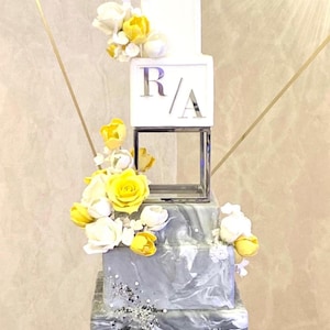 Peut inclure: Un gâteau de mariage à quatre étages avec un étage inférieur à motif marbré. Le deuxième étage est blanc avec les initiales "R/A" en lettres argentées. Les deux étages supérieurs sont blancs et décorés de fleurs en sucre jaunes et blanches.
