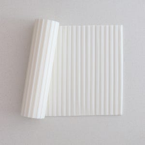 Puede incluir: Un objeto rectangular blanco texturizado con ranuras verticales, parcialmente enrollado. El objeto está sobre una superficie gris claro. Las ranuras crean un patrón de luz y sombra, dándole una apariencia tridimensional. La estética general es limpia y minimalista.