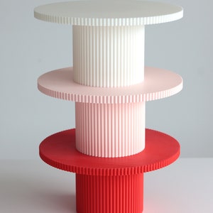 Puede incluir: Un soporte para tartas de tres niveles con niveles blancos, rosas y rojos. Cada nivel tiene un borde acanalado y una base cilíndrica.
