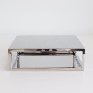 May include: A silver two-tiered table with a mirrored bottom shelf. The table is made of polished stainless steel and has a modern, minimalist design.