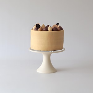 Puede incluir: Un pastel de un solo nivel con glaseado marrón claro y detalles de chocolate y caramelo. El pastel está sobre un soporte para pastel blanco.