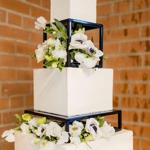 Może przedstawiać: Czteropiętrowy biały tort ślubny z czarnymi kwadratowymi podstawkami na torty ozdobionymi białymi kwiatami.