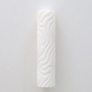 May include: A white, cylindrical candle with a textured surface featuring a flowing, wave-like design. The candle stands vertically against a plain white backdrop. The pattern gives the candle a dynamic appearance, enhancing its simple shape.