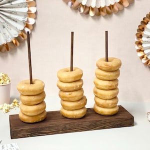 Pode incluir: Um suporte de madeira para donuts com três camadas de donuts glaciados. O suporte é de madeira escura e tem um visual rústico. Os donuts estão empilhados em pinos de madeira e estão prontos para serem apreciados.