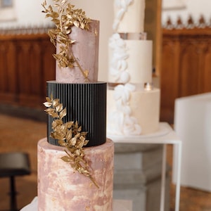 Könnte beinhalten: Eine dreistöckige Hochzeitstorte mit einer schwarz-weiß gestreiften Etage, einer rosafarbenen Etage mit goldenen Blattverzierungen und einer unteren Etage mit einer marmorierten rosa-goldenen Oberfläche. Die Torte ist mit goldenen Blattzweigen verziert.