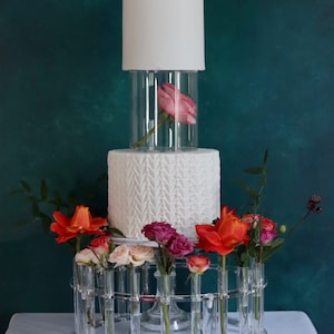 May include: A white cake with a delicate floral design is displayed on a clear acrylic cake stand. The cake is topped with a clear cylinder containing a single pink rose. The stand features multiple clear glass tubes that hold a variety of colorful flowers, including red, pink, and peach.