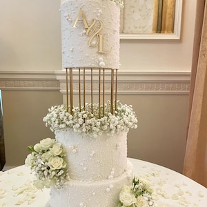 Könnte beinhalten: Eine dreistöckige weiße Hochzeitstorte, verziert mit weißen Perlenstreuseln und Gipskrautblüten. Die oberste Etage hat einen goldenen Ständer mit goldenen Buchstaben "A & L" auf der Torte.