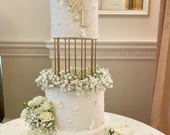 May include: A three-tiered white wedding cake decorated with white pearl sprinkles and baby's breath flowers. The top tier has a gold stand with gold letters "A & L" on the cake.