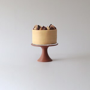 Puede incluir: Un pequeño pastel redondo con un glaseado marrón claro y decoraciones de chocolate y caramelo en la parte superior. El pastel está en un soporte para pastel marrón.