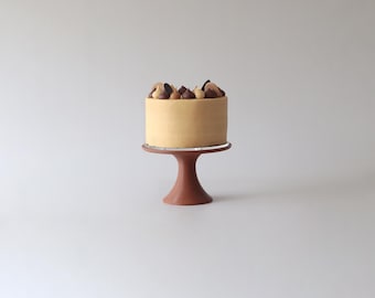 May include: A small, round cake with a light brown frosting and chocolate and caramel decorations on top. The cake is on a brown cake stand.
