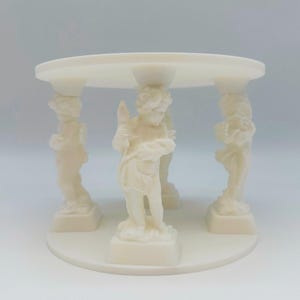 Könnte beinhalten: Ein cremefarbener dekorativer Ständer mit vier Cherub-Statuen, die eine runde Plattform tragen. Die Cherubim befinden sich in verschiedenen Posen und halten Gegenstände. Der Ständer hat eine kreisförmige Basis und besteht aus einem glatten, festen Material.