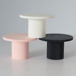 Könnte beinhalten: Drei dreistufige Tortenständer in Weiß, Rosa und Schwarz. Jeder Ständer hat eine runde Oberseite und eine geriffelte Basis.