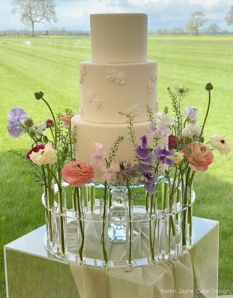 May include: A three-tiered white wedding cake with delicate floral designs, displayed on a clear acrylic stand with a circular arrangement of glass tubes holding colorful flowers.