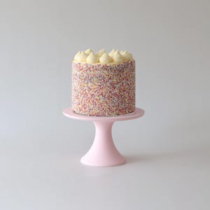 Könnte beinhalten: Eine einstufige Torte, die mit bunten Streuseln und weißem Frosting verziert ist, auf einem rosa Tortenständer.