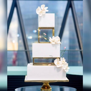 Peut inclure: Un gâteau de mariage blanc à quatre étages avec des cadres géométriques dorés et des accents de fleurs d'orchidées blanches.