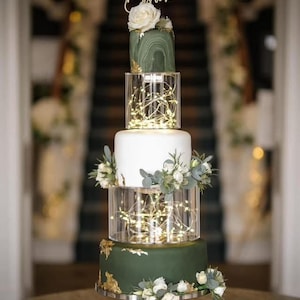 Könnte beinhalten: Eine vierstöckige Hochzeitstorte mit grünem und weißem Frosting. Die oberste Schicht ist mit einem grünen Marmormuster und einem Holzschild verziert, auf dem "Mr. & Mrs. Brown" steht. Die Torte ist mit weißen Blumen und Grün verziert. Die Torte steht auf einem silbernen Tortenständer.