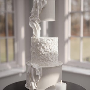 Puede incluir: Una tarta de boda blanca de tres pisos con intrincados diseños florales y adornos de tela. La capa superior parece flotar sobre la capa media. Una vela blanca encendida está sobre una mesa negra.