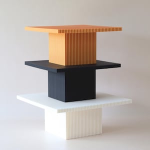 Może przedstawiać: Trzy piętrowe podstawki na tort, ułożone jedna na drugiej. Górna podstawka jest pomarańczowa, środkowa jest czarna, a dolna jest biała. Każda podstawka ma kwadratowy blat i kwadratową podstawę z pionowymi rowkami.