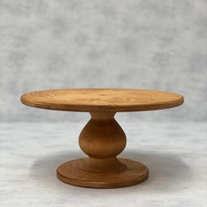 Könnte beinhalten: Ein Kuchenständer aus Holz mit runder Oberseite und einem Sockel. Der Ständer ist aus Naturholz gefertigt und hat eine hellbraune Farbe.