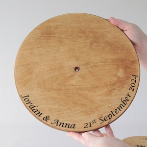 May include: A round, light-brown wooden disc with engraved text. The text reads "Jordan & Anna 21st September 2024". The disc has a small hole in the center and is held by two hands. The item appears to be a wedding keepsake.