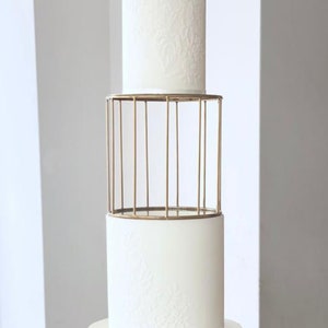 Peut inclure: Un gâteau de mariage blanc à trois étages avec des motifs en relief complexes. Le niveau du milieu est sur un support en métal doré avec un design en forme de cage.