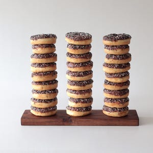 Pode incluir: Três pilhas de donuts glaciados de chocolate com granulado numa tábua de madeira. Cada pilha tem 10 donuts.