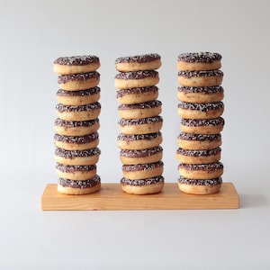 Puede incluir: Tres pilas de donuts glaseados con chocolate y espolvoreados, cada pila tiene aproximadamente 10 donuts de altura, sobre una tabla de madera.