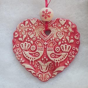 May include: A heart-shaped, red ceramic ornament with a detailed, raised white design featuring birds, flowers, and other decorative elements. A small wooden bead with a snowflake design and a red ribbon are attached for hanging.