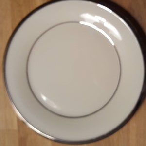 May include: A white ceramic plate with a silver rim. The plate is round and has a smooth surface.