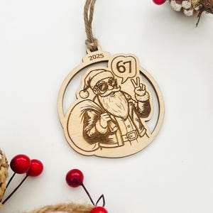 May include: Wooden Christmas ornament featuring Santa Claus wearing sunglasses and giving a peace sign. The ornament has the number "67" in a speech bubble and the year "2025". The ornament is surrounded by pine cones and red berries.