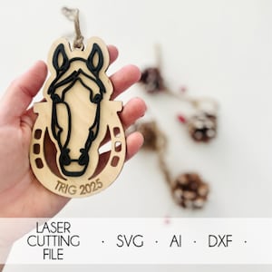 Pode incluir: Um ornamento de cabeça de cavalo de madeira com uma base de ferradura. O rosto do cavalo é delineado em preto, com o texto "TRIG 2025" abaixo. O ornamento é segurado em uma mão, com outros itens decorativos no fundo. A parte inferior da imagem diz "LASER CUTTING FILE".