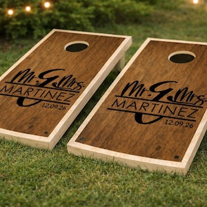 Puede incluir: Dos tableros de cornhole de madera con tinte oscuro y borde de madera clara. Cada tablero tiene un agujero central y presenta el texto "Mr. & Mrs. MARTINEZ" y la fecha "12.09.26" en negro. Los tableros est&aacute;n sobre hierba verde.