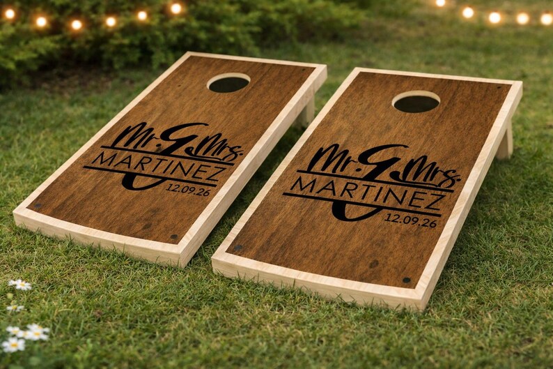 Puede incluir: Dos tableros de cornhole de madera con tinte oscuro y borde de madera clara. Cada tablero tiene un agujero central y presenta el texto "Mr. & Mrs. MARTINEZ" y la fecha "12.09.26" en negro. Los tableros est&aacute;n sobre hierba verde.