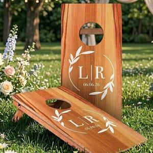 Puede incluir: Dos tableros de cornhole de madera con un acabado de vetas naturales. Cada tablero presenta un agujero circular y letras blancas que dicen "LR" con la fecha "06.06.26" dentro de una corona decorativa. Un tablero est&aacute; en posici&oacute;n vertical, el otro est&aacute; en &aacute;ngulo.