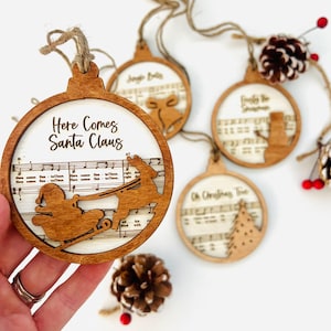 May include: Wooden Christmas ornaments with festive designs. One ornament features Santa Claus in a sleigh with the text "Here Comes Santa Claus." Other ornaments include a bell, snowman, and Christmas tree. Each ornament has a musical score background.